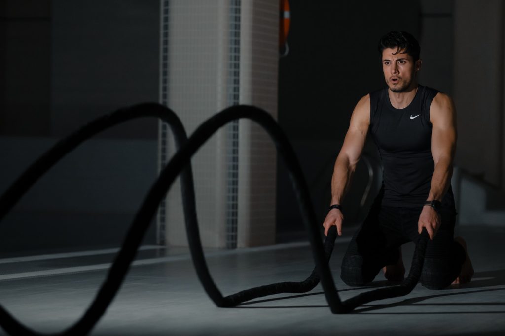 Man in gym workout with battle ropes wearing black athletic gear kneeling on the floor focused on exercise fitness training