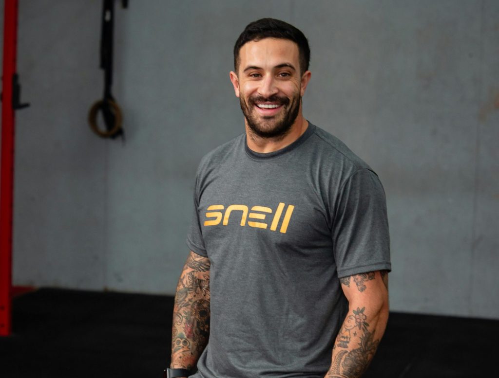 Athletic man with tattoos smiling in a gym wearing sportswear.